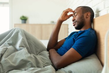 African American Man Having Headache Touching Forehead Suffering From Pain With Eyes Closed Sitting In Bed In Modern Bedroom At Home. Male Health Problem, Migraine Symptom, Side View