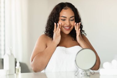 Facelift and skin care concept. Young black plus size woman massaging her face and looking at mirror. African american lady applying moisturizing cream or doing facial anti aging massage