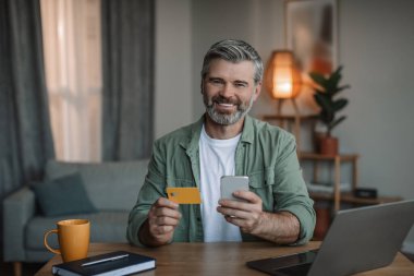 Buy and pay at home. Smiling retired european man with beard uses smartphone, computer and credit card, shopping online in room interior. Sale, business and offer with device during covid-19 outbreak