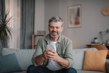 Glad elderly european man with beard chatting on smartphone, watching video, playing game in living room interior. Free time for blog at home, new app, advertisement and offer during covid-19 outbreak