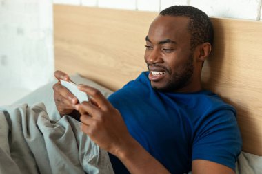 Smiling African American Male Using Mobile Phone Playing Game Online Lying In Bed At Home. Cheerful Man Texting Via Cellphone In Bedroom. Technology Concept. Selective Focus