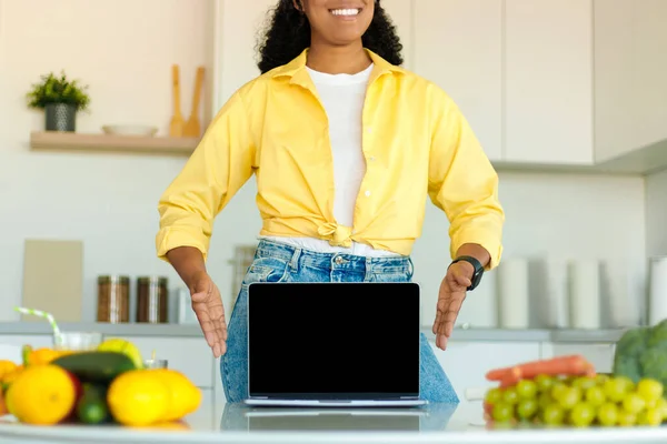 African american lady showing laptop with blank screen at table with organic vegetables at kitchen interior, mockup. Cooking dinner at home, new website, recipe, ad