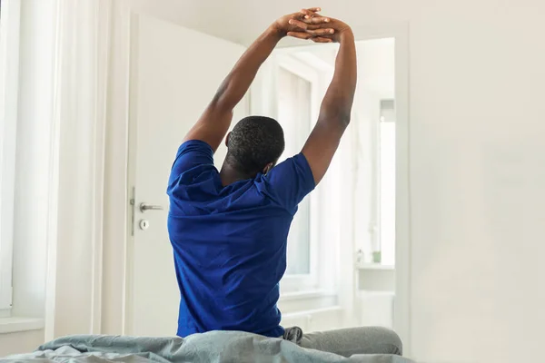 Back View Shot Of African American Young Man Stretching Arms Sitting On Bed In Modern Bedroom At Home, Wearing Blue T-Shirt. Good Morning, Weekend Concept