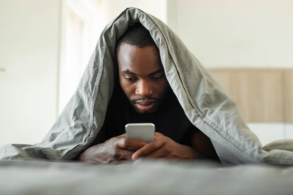 Sleepy African American Man Using Cellphone Texting Lying Covered Blanket — Fotografia de Stock