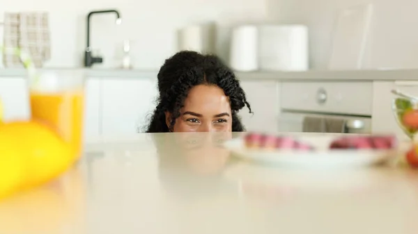 Healthy or unhealthy food. Positive black woman choosing between fresh fruit juice and donuts, peeking out of table in kitchen interior