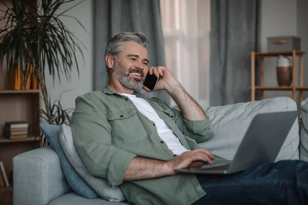Glad mature european man with beard calls by phone, works on laptop, sits on sofa in living room interior. Ad, offer remote at home, modern technology, business and new normal during covid-19 outbreak