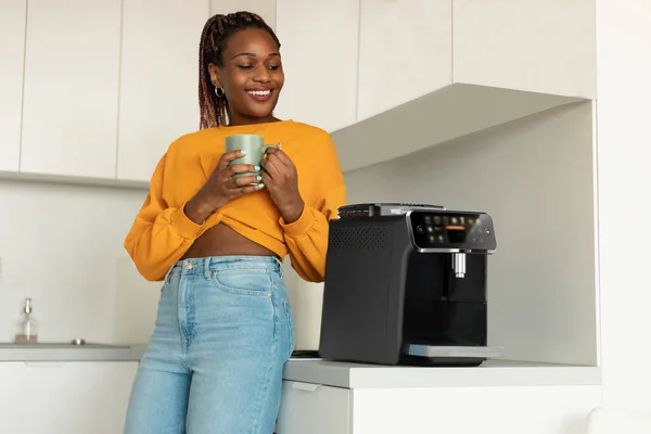 Happy african american woman enjoying fresh aromatic coffee near modern machine in kitchen interior, drinking morning hot beverage at home