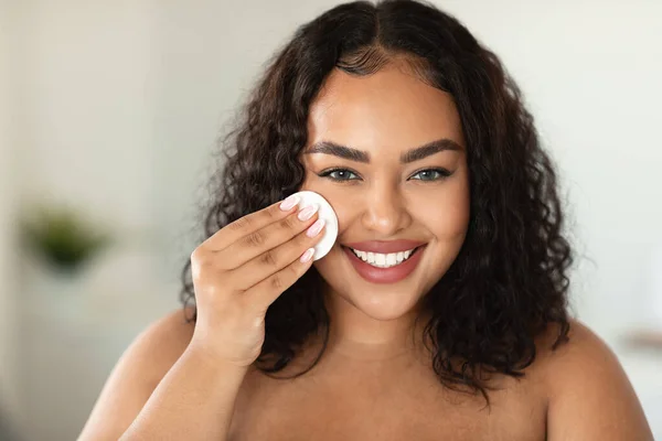 Makeup remover, cleansing and routine procedures. Happy african american oversize woman wiping her face with cotton sponge and smiling at camera