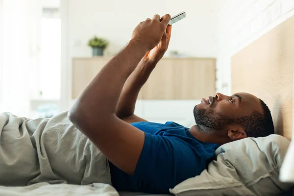 African American Guy Using Phone Lying On Back, Reading Messages Texting In Modern Bedroom At Home. Male Using Mobile Application On Smartphone Resting In Bed In The Morning. Side View