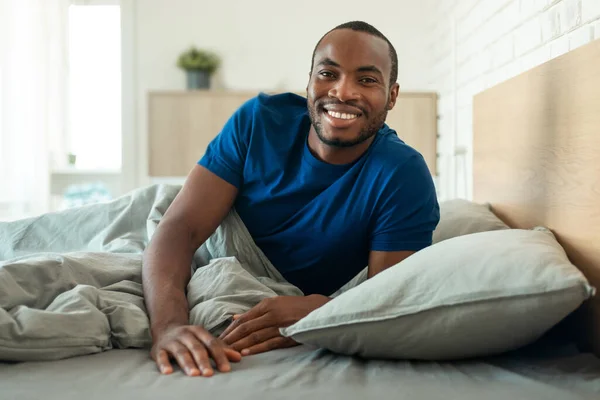 Happy African American Man Smiling To Camera Lying In Bed At Home In The Morning. Well-Slept Guy Posing In Modern Bedroom Indoor. Comfort And Sleeping Routine Concept