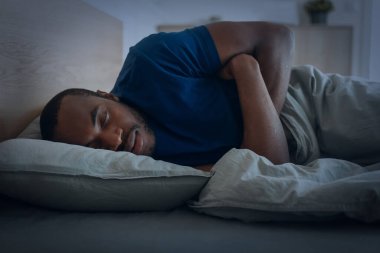 Black Guy Sleeping Lying Under Blanket In Bedroom At Home At Night. Male Resting Napping Peacefully In Bed. Recreation And Relaxation Concept. Low Light