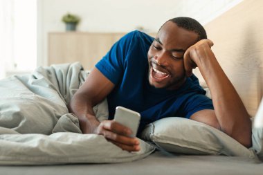 Cheerful Black Man Using Smartphone Browsing Internet Having Fun Lying In Bed In Modern Bedroom At Home On Weekend. Technology And Gadgets, Mobile Communication Concept