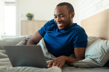 African American Man Using Laptop Computer Working Online Lying In Bed At Home. Male Freelancer Browsing Internet Doing Work Remotely In Modern Bedroom. Technology Concept