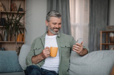 Glad retired european man with beard chatting on smartphone, enjoying drink and chat in social networks in living room interior. Ad, offer and new app at home alone in spare time due covid-19 outbreak
