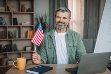 Cheerful retired european man with beard teacher hold USA flag shooting video lesson on laptop in room interior. Learn english language, tutor work at home, study and education due covid-19 outbreak