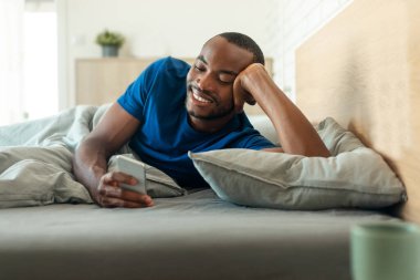 Cheerful African American Guy Using Cellphone Lying In Bed In Bedroom At Home. Gadgets And Technology, Mobile Communication And Gadgets Concept. Selective Focus
