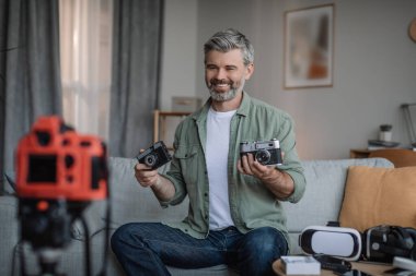 Cheerful elderly caucasian man with beard shooting video for blog, making review on photo cameras in room interior. Recommendation, offer and ad, hobby , modern work remotely at home due covid-19