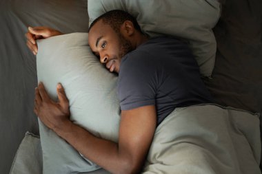 Sleeplessness. Black Millennial Guy Suffering From Insomnia Lying In Bed Hugging Pillow In Modern Bedroom At Home. Sleeping Routine Problems Concept. Above View Shot