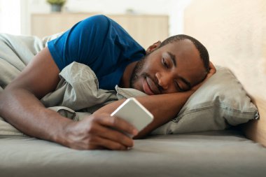 African American Millennial Guy Using Cellphone Lying Covered With Blanket In Bed At Home. Male Using New Application And Browsing Internet On Smartphone. Mobile App And Gadgets. Selective Focus