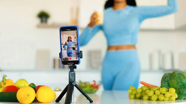 Excited fit black woman recording new video for her sport and food blog on smartphone, standing in kitchen and showing biceps at cellphone camera, selective focus