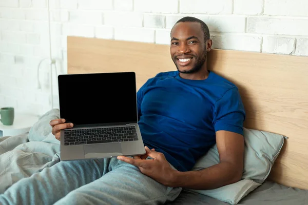 Black Man Showing Laptop Computer With Blank Screen Working Online Sitting In Bed At Home. Male Freelancer Recommending Great Website In Modern Bedroom. Mockup