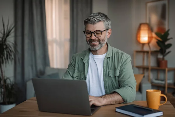Cheerful elderly european man with beard in glasses watches video lesson, chatting on computer in living room interior. Freelance, business and work at home with modern device during covid-19 outbreak