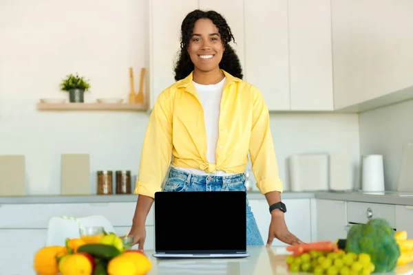 Cooking recipes online. Happy black lady showing laptop with blank screen in modern kitchen, mockup, free copy space. Space for your advertisement