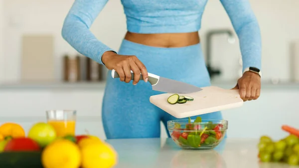 Detox recipes. Fit black lady cooking healthy vegetable salad, cutting cucumber on wooden board, standing in modern kitchen, cropped. Young woman preparing dinner for weight loss dieting