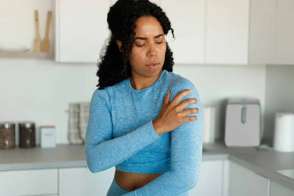 Sporty black woman putting her hand on injury shoulder, having pain, standing in kitchen interior at home, resting after domestic training. Sport concept