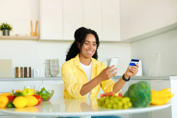 Ordering food online. Young african american woman with smartphone and credit card purchasing grocery delivery from internet, sitting in kitchen interior