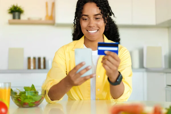 Cheerful black woman buying food online, using cellphone and plastic credit card, purchasing ingredients for healthy dinner, cooking for family at home, copy space