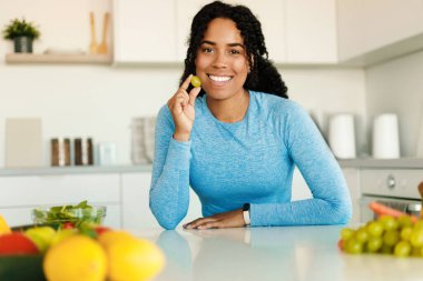 Excited fit african american lady eating fruits for breakfast, holding grape and smiling at camera, sitting at table in kitchen, free copy space