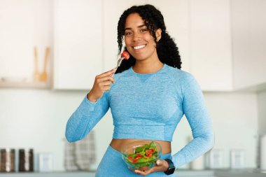 Healthy dieting. Positive black fit woman holding fork and bowl with fresh vegetable salad, smiling at camera, standing in kitchen. Portrait of sporty lady having fresh balanced meal