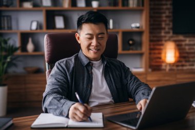 Cheerful mature korean male working at table with laptop, businessman analyzing data stock in home office interior. Trading, marketing, digital technology, app and solutions for business development