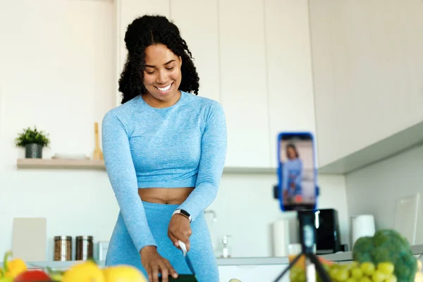 Happy african american lady in sportswear recording video on smartphone camera, preparing fresh salad in kitchen. Woman cutting vegetables on table