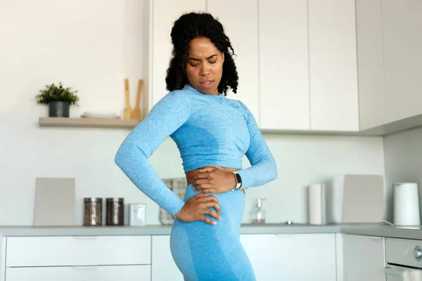 Dissatisfied fit black lady holding aching hip, has kidney inflammation, touching location of paim, wearing sport clothes, having sporty shaped body