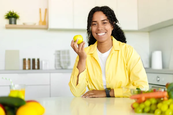 Weight loss diet. Happy black lady holding apple and smiling at camera, sitting in modern kitchen interior, copy space. Healthy eating and nutrition for slimming