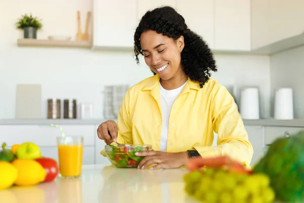 Happy black lady eating fresh vegetable salad, enjoying healthy food and drink, sitting in modern kitchen at home. Nutrition and weight loss concept
