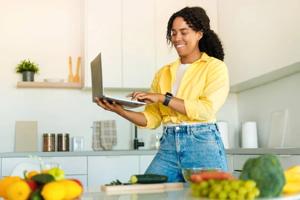 Food blog about cooking. Happy african american woman typing on laptop, preparing healthy meal on table in kitchen. Lady searching for new recipes