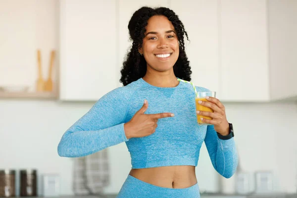 Happy african american lady in sportswear pointing finger at glass of fresh orange juice, standing in kitchen and smiling at camera, copy space. Vitamins, diet and weight loss