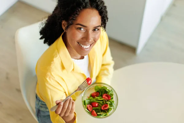 Happy african american lady eating salad with organic vegetables and smiling at camera, sitting at table, above view. Food blog, cooking proper nutrition for health care