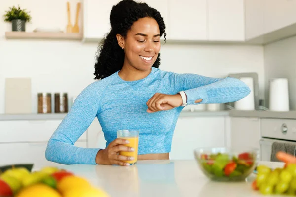Healthy lifestyle. Happy fit african american woman in sports bra holding fresh juice and looking at wrist smartwatch, sitting at table in kitchen