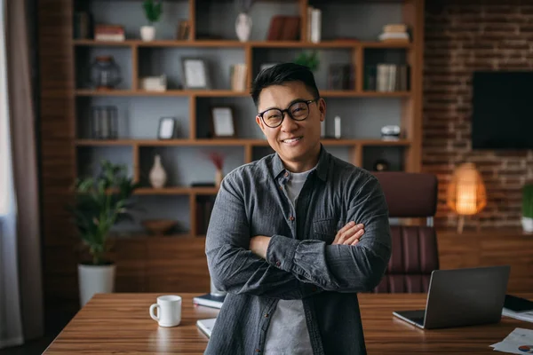 Smiling confident successful adult chinese man in glasses with crossed arms on chest looking at camera in office interior. Businessman at work at home, development and business, trading and marketing