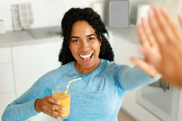 Happy african american fit lady drinking fresh juice and giving high five to friend, standing in kitchen interior and smiling. Positive woman in sportswear resting after domestic workout