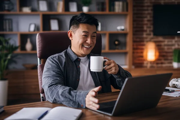 Glad adult chinese man ceo manager work with laptop with cup of drink and talking with client in home office interior. Businessman have video call and coffee break, development and social distance