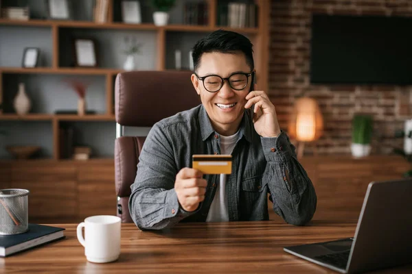 Glad middle aged korean man in glasses calls by phone use laptop and credit card, shopping online in office interior. Professional working new start up, credit and finance, businessman enjoy sale