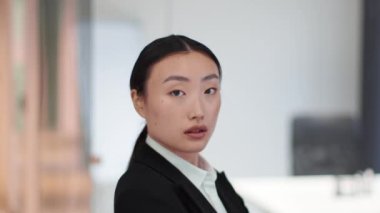 Professional young confident asian business lady turning face to camera and looking seriously, standing at office interior, side view, slow motion