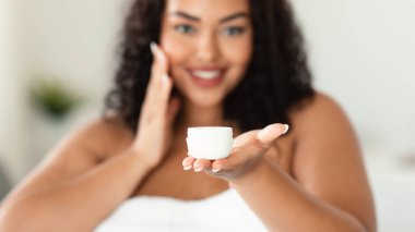 African american plus size woman holding jar of organic skin cream, applying moisturiser on face after shower, selective focus, cropped, panorama