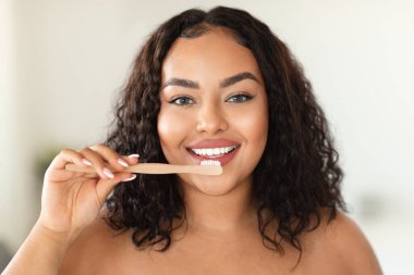 Healthy teeth and care. Happy black plus size lady brushing teeth with toothbrush and smiling at camera in bathroom interior in the morning, closeup