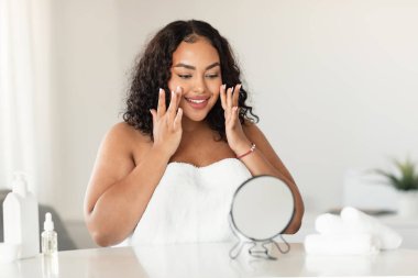 Skincare concept. Happy black oversize woman touching her face while looking at mirror in bedroom, enjoying skin condition after beauty treatments, copy space
